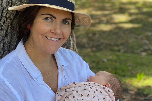 Kate wearing a sunhat and white shirt, holding baby Ava in pink swaddle, leaning against a tree in a park.